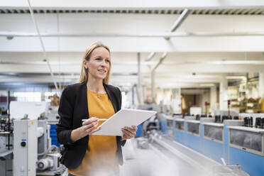 Lächelnde blonde Geschäftsfrau mit Tablet-PC in einer Fabrik stehend - DIGF17575