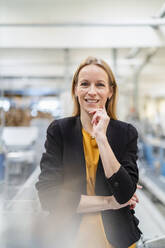 Happy blond businesswoman standing with hand on chin in factory - DIGF17580