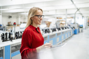 Glückliche Geschäftsfrau mit Brille hält Tablet-PC in einer Fabrik - DIGF17631