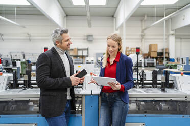 Lächelnder Geschäftsmann im Gespräch mit einem Kollegen, der einen Tablet-PC an einer Maschine in einer Fabrik benutzt - DIGF17638