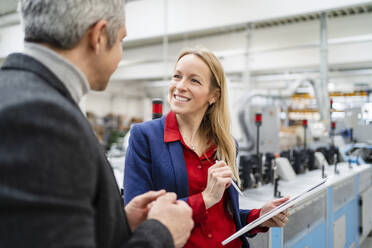 Lächelnde blonde Geschäftsfrau mit Tablet-PC und Blick auf einen Kollegen, der in einer Fabrik diskutiert - DIGF17643