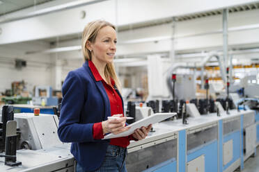 Lächelnde blonde Geschäftsfrau mit Tablet-PC an einer Maschine in einer Fabrik - DIGF17644