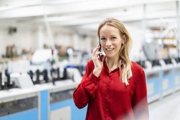 Lächelnde blonde Geschäftsfrau, die in einer Fabrik mit einem Smartphone telefoniert - DIGF17650