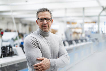 Smiling businessman wearing eyeglasses standing with arms crossed in factory - DIGF17662