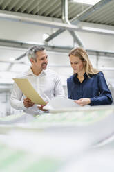 Smiling businessman with documents looking at blond businesswoman reading reports in factory - DIGF17675