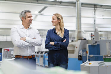 Smiling businesswoman and businessman standing with arms crossed looking at each other in factory - DIGF17678