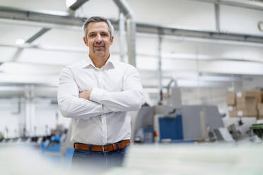 Smiling businessman standing with arms crossed in factory - DIGF17681