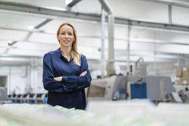 Smiling blond businesswoman standing with arms crossed in factory - DIGF17684