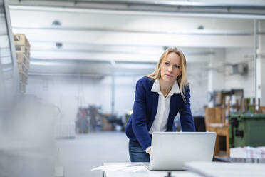 Nachdenkliche blonde Geschäftsfrau mit Laptop, die sich auf einen Schreibtisch in einer Fabrik lehnt - DIGF17704