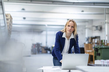Thoughtful blond businesswoman with laptop leaning on desk in factory - DIGF17704