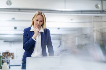 Blond businesswoman with hand on chin looking at laptop in factory - DIGF17705