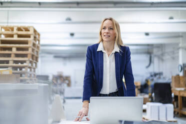 Smiling businesswoman with laptop standing at desk in factory - DIGF17706