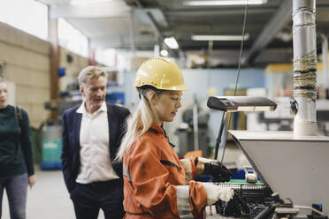 Female industrial worker in hardhat working on machinery while managers in background at factory - MASF28236