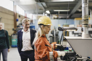 Weiblicher Industriearbeiter mit Schutzhelm bei der Arbeit an Maschinen, während im Hintergrund Manager in einer Fabrik zu sehen sind - MASF28236