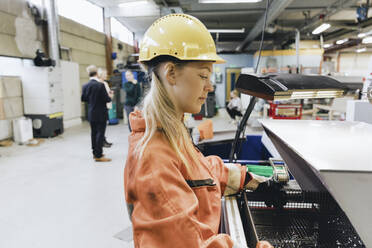 Seitenansicht einer Arbeiterin mit Schutzhelm, die in einer Fabrik Maschinen bedient - MASF28244