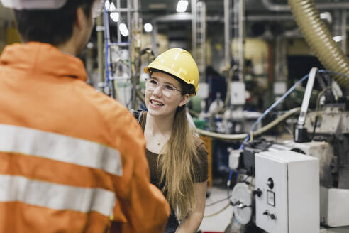 Lächelnde Managerin mit Schutzhelm im Gespräch mit einem männlichen Arbeiter in einer Fabrik - MASF28253