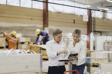 Businesswoman and businessman discussing over digital tablet while workers working in background at warehouse - MASF28259