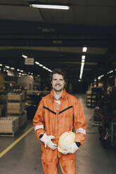 Portrait of smiling male worker in protective workwear standing in warehouse - MASF28272