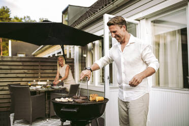 Lächelnder reifer Mann, der eine Grillmahlzeit zubereitet, während eine Frau im Hintergrund im Hinterhof steht - MASF28293