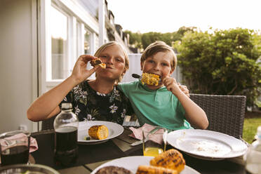 Porträt von Geschwistern, die gegrillten Mais und Fleisch essen, während sie im Hinterhof sitzen - MASF28295
