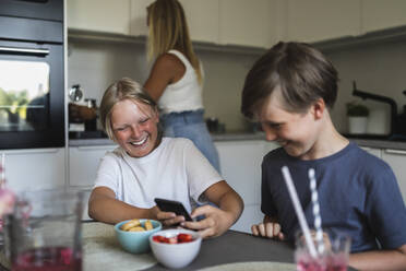 Lächelnde Brüder, die ein Smartphone benutzen, während die Mutter im Hintergrund Küchenarbeit macht - MASF28304