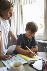Smiling boy standing by brother doing homework at home - MASF28307