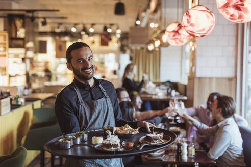 Porträt eines männlichen Kellners mit einem Essenstablett, während die Kunden im Hintergrund an der Bar anstoßen - MASF28335
