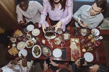 Frau, die mit ihrem Smartphone fotografiert, während sie mit Freunden im Restaurant zu Abend isst - MASF28344