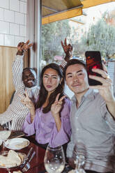 Männliche und weibliche Freunde machen ein Friedenszeichen, während sie ein Selfie mit ihrem Smartphone in einer Bar machen - MASF28347