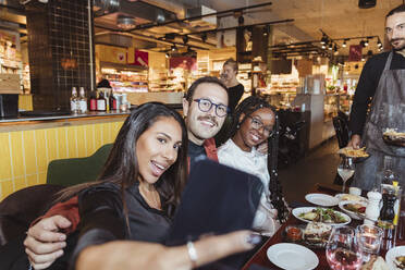 Cheerful woman taking selfie through smart phone with male and female friends in restaurant - MASF28351