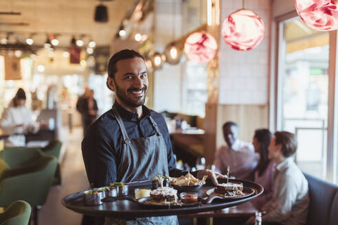 Portrait of smiling waiter with food while customer sitting in background at bar - MASF28412