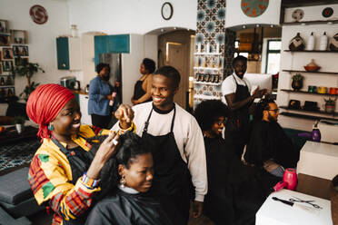 Lächelnde Friseurin im Gespräch mit einem männlichen Mitarbeiter, während sie einem Kunden im Friseursalon die Haare schneidet - MASF28431