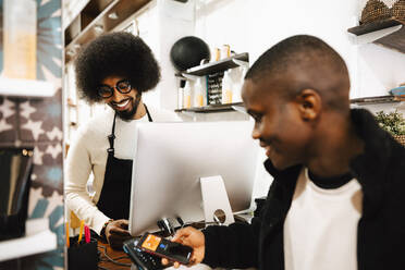 Smiling male customer doing contactless payment through smart phone at barber shop - MASF28473