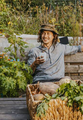 Lächelnder Landwirt mit Kaffeetasse auf einer Bank auf einem Bauernhof - MASF28553