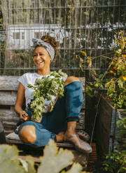 Cheerful female farmer looking away while sitting on bench at community garden - MASF28600