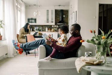 Smiling father teaching daughter sitting on chair in living room - MASF28634