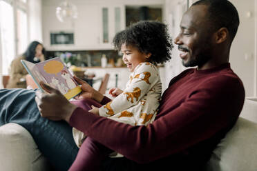 Glücklicher Vater liest mit seiner Tochter ein Buch im Wohnzimmer zu Hause - MASF28635