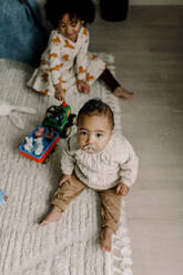 Männliches Kleinkind mit Schnuller auf dem Teppich sitzend, während die Schwester im Hintergrund zu Hause ist - MASF28642