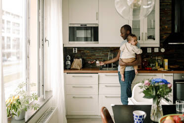 Vater mit Babyflasche trägt männliches Kleinkind in der Küche zu Hause - MASF28665