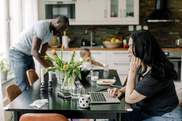 Businesswoman freelancing on laptop while family in background at home - MASF28687