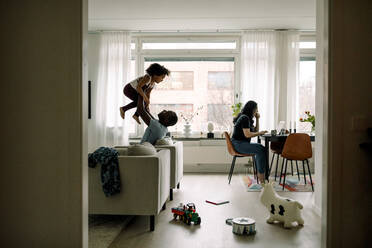 Father carrying daughter while mother freelancing on laptop in living room - MASF28692