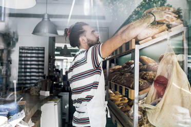 Junger männlicher Unternehmer beim Anrichten von Lebensmitteln durch das Glas einer Bäckerei gesehen - MASF28787