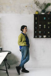 Lächelnde Besitzerin mit Hand in der Tasche, die sich an die Wand einer Werkstatt lehnt - MASF28857
