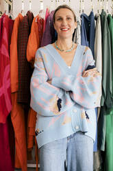 Portrait of female owner with arms crossed standing against clothes in boutique - MASF28873