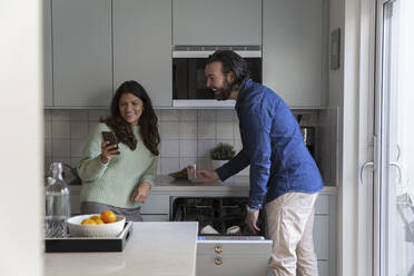 Smiling woman sharing smart phone with man in kitchen - MASF28883