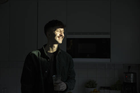 Lächelnder Mann mit Sonnenlicht im Gesicht in der Küche stehend - MASF28917