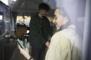 Smiling woman using smart phone standing by boyfriend seen through glass window - MASF28940