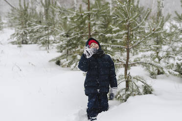 Glücklicher Junge in warmer Kleidung im verschneiten Wald an einem Baum stehend - SEAF00517