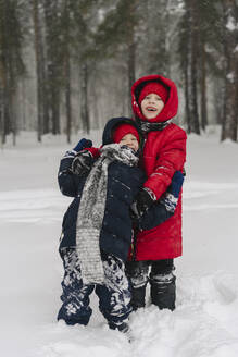 Smiling boy embracing brother standing on snow in forest - SEAF00540