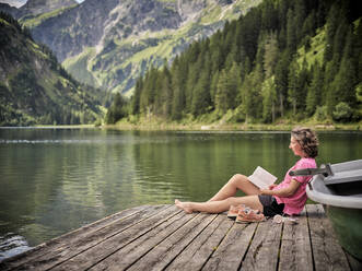 Woman reading book by rowboat at lake - DIKF00646